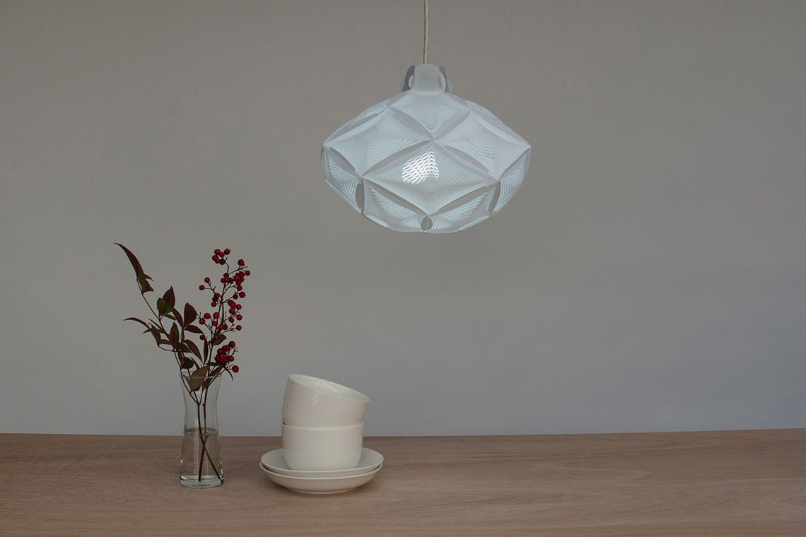 Airy RI-20 white geometric pendant lamp in the shape of rhombic icosahedron is hanging over a wood dining table with a glass vase and white cups
