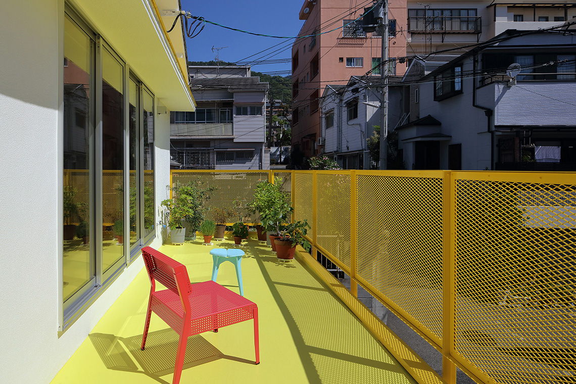 24d-studio 2nd floor bright yellow east balcony with plants and pink and blue chairs in the background